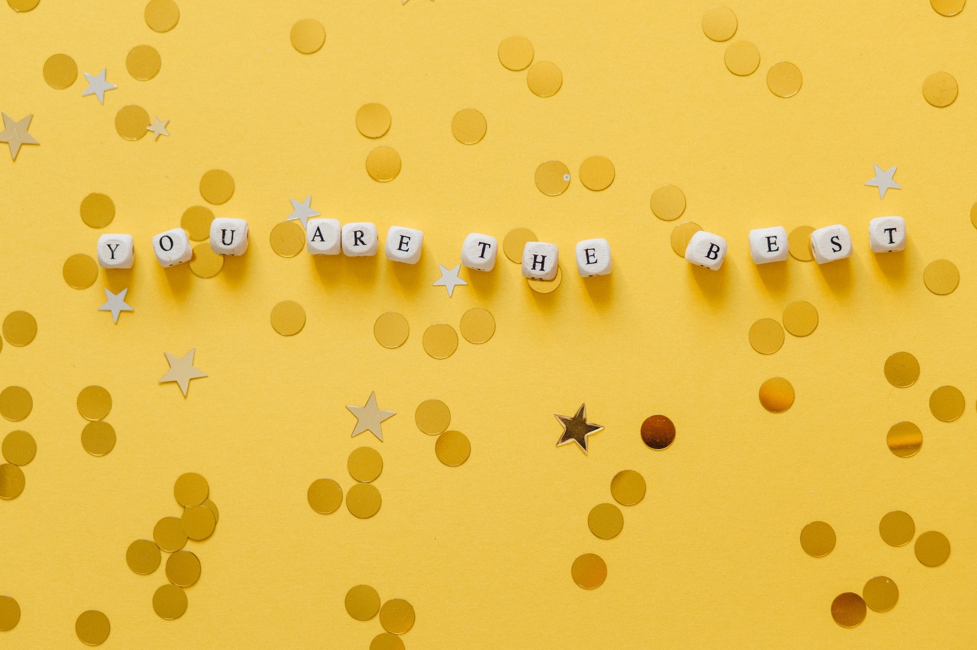 Auf einer gelben Oberfläche liegen mehrere Würel mit Buchstaben bedruckt. Zusammen bilden die Würfel den Satz "You are the best". Drumherum ist goldfarbenes Konfetti verteilt.