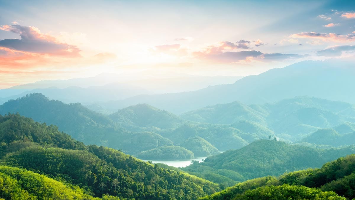 Naturfotografie. Blick von einem Berg auf eine bergige bzw. hügelige Landschaft, dicht und grün bewaldet. Hinter ein paar Wolken strahlt die Sonne in Gelb- und Rosatönen.
