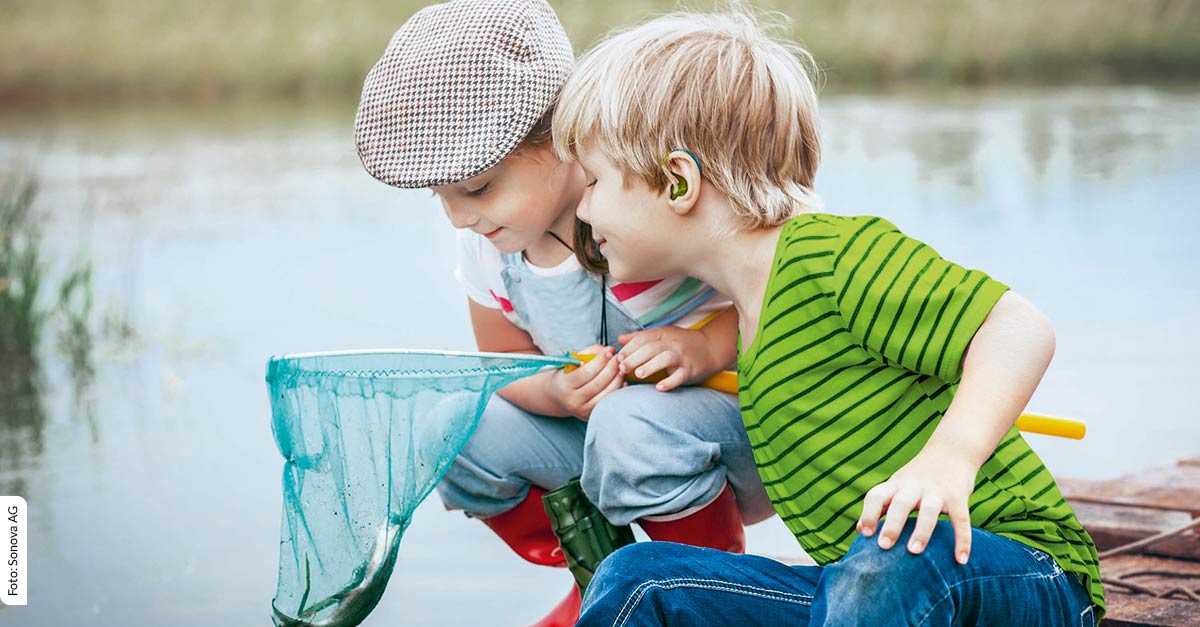 Zwei Kinder am Teich, die gerade einen Fisch im Netz gefangen haben. Eines der Kinder trägt eine Hörlösung von Hidden Champion Sonova